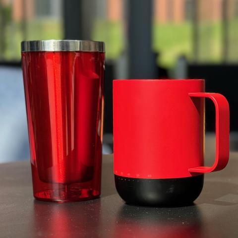 A red reusable tumbler and a red and black reusable mug sit on a cafe table.
